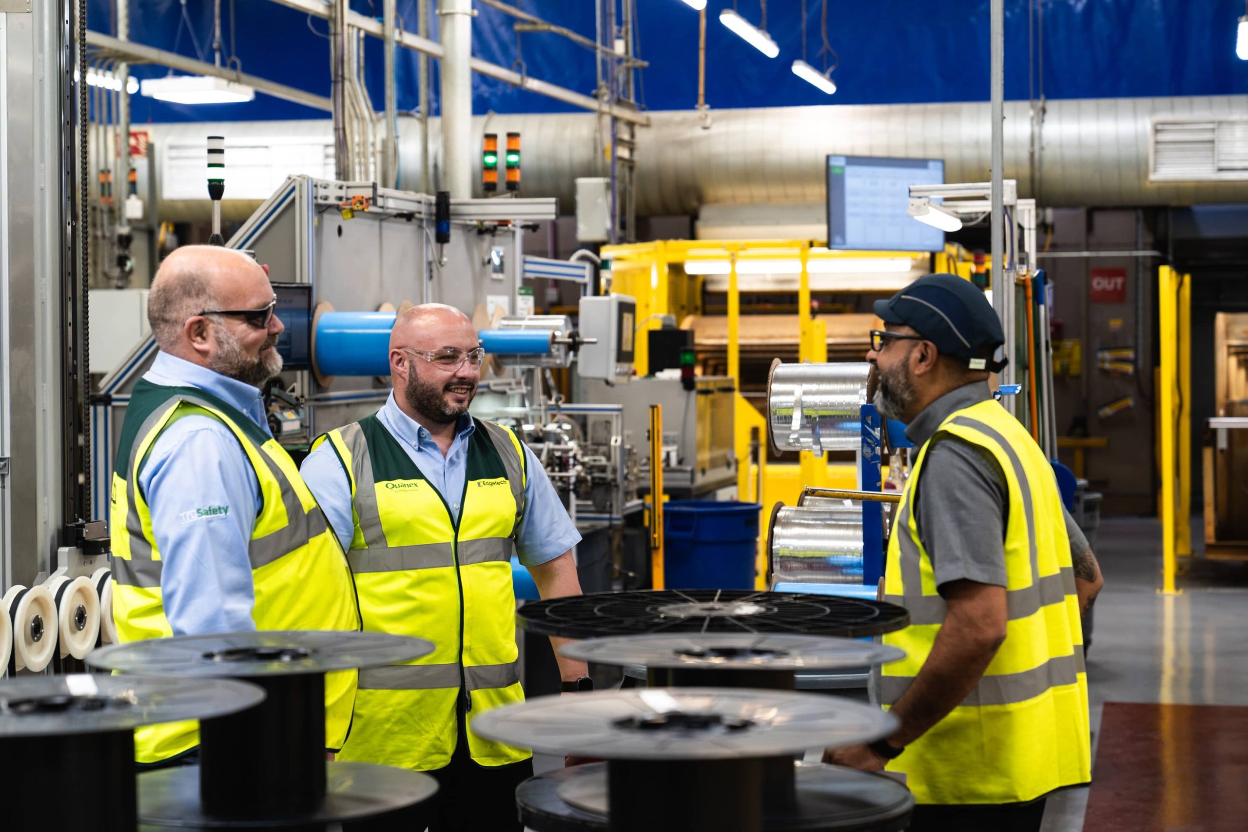 Three men in safety vests at an Edgetech factory, reviewing plans while surrounded by machinery and safety signage.